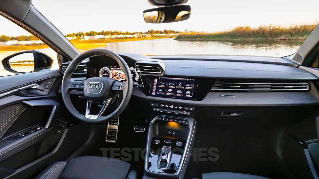 El elegante interior del Nuevo Audi A3 2025 cuenta con modernas pantallas digitales, un volante de última generación y una sofisticada consola central, todo ello enmarcando perfectamente una vista serena del lago a través del parabrisas. - ofertamotor.es