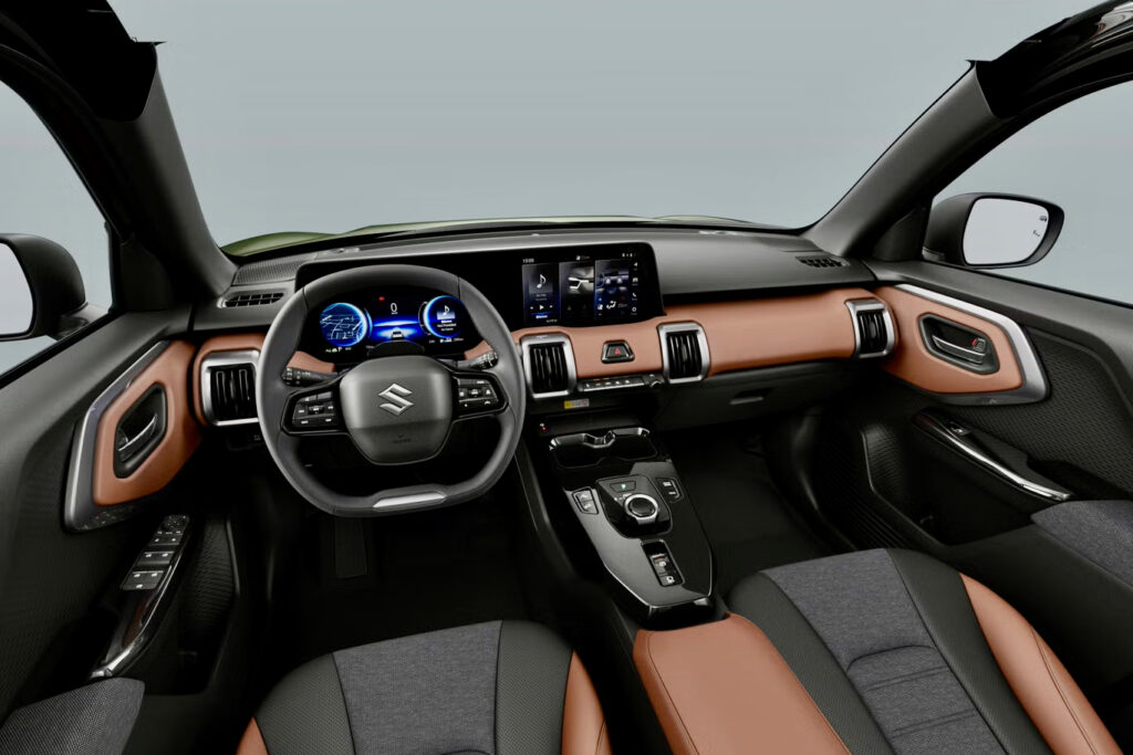 El interior del Suzuki e Vitara presenta un elegante volante con el logotipo icónico, complementado por pantallas digitales y una elegante combinación de tapicería marrón y negra.